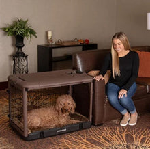 a happy woman sitting on a leather couch next to her dog inside a brown dog steel crate with bolster pad in the modern living room with book shelf and flower pot on the back 