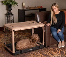a happy woman sitting on a leather couch staring at her dog laying inside a tan colored steel dog crate and a flower pot on the background 