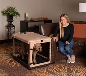 a happy woman sitting on a leather couch next to her dog inside a tan colored steel dog crate holding the steel gate of the crate open for her dog in a modern living room setting 