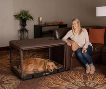 a happy woman sitting on a leather couch starring at her dog inside a large brown dog steel crate laying on a bolster pad in a modern living room with bookshelf at the back and flower pot 