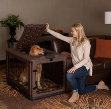 a happy woman kneeling on the floor holding the top door open for her dog inside a large brown steel dog crate next to a leather couch in a modern room with bookshelf and flower pot 