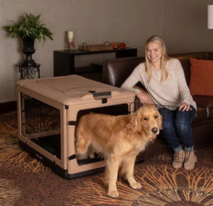 a happy woman sitting on a leather couch holding the steel door open for her dog to get out of a large tan dog steel crate in a modern living room with bookshelf at the back and flower pot