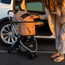 a lady poking her hands on a dessert rose colored dog stroller next to a white car on the road 