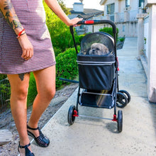 a tattooed lady holding on a black dog stroller with red buttons on the handle bars and a cute dog in it  at the edge of a street with green bushes on the other side