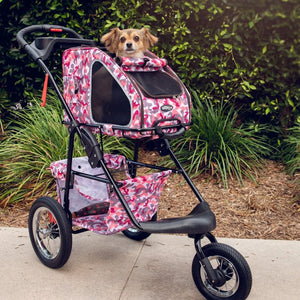 a cute dog riding a pink camo dog stroller inside a dog carrier next to some tall grasses