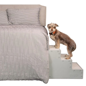 a fluffy tiny dog standing on a grey colored three step dog stair next to a white bed 