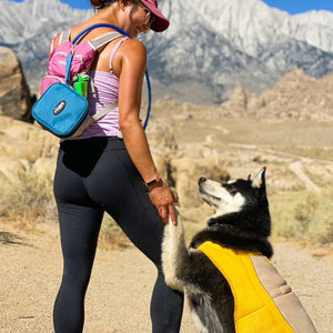 a woman holding her dogs hand hiking with a bag of portable dog bowl on her back 