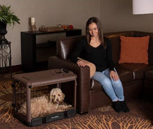 a woman siting on a leather couch staring ate her dog inside a brown colored steel dog crate with bolster pad in a modern living room with wooden shelf and a pot of flower at the back
