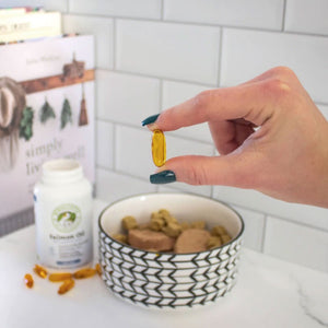 a hand of a woman holding a capsule gel of a dog supplement next to a bowl of dog food and books
