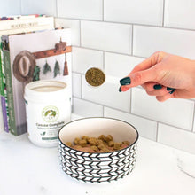 A picture of a lady's hand showing a scoop of the Canine Complete Multivitamins next to a feeding bowl with dog food and some books