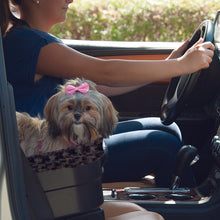 A close up image of a shih-tzu on a brown leather car seat inside a Bucket Seat Dog Booster with Chocolate Insert, 17" next to a driving lady