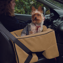 Close up image of a yorki sitting inside a large car booster in tan color next to a driving lady in black 