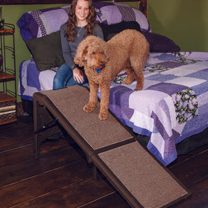 a lady sitting on the bed with her golden doodle getting off the bed through a chocolate colored dog ramp in their bedroom