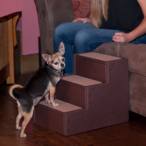 a chihuahua standing on the first step of a chocolate colored pet stair next to a lady sitting on a brown couch